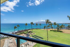 A view of a pool area with lounge chairs and a pool, a grassy lawn with palm trees, and a beach with the ocean and a mountain in the background. - Kihei Surfside #508: Gorgeous Oceanfront Escape with Mesmerizing Views (Kihei, Maui)