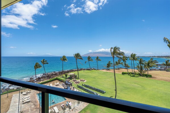 A view of a pool area with lounge chairs and a pool, a grassy lawn with palm trees, and a beach with the ocean and a mountain in the background. - Kihei Surfside #508: Gorgeous Oceanfront Escape with Mesmerizing Views (Kihei, Maui)