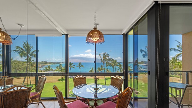 A dining area with a glass table and wicker chairs, overlooking a beach and palm trees.