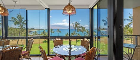 A dining area with a glass table and wicker chairs, overlooking a beach and palm trees.
