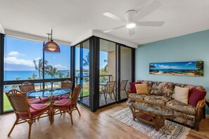 Un vaste espace de vie avec un balcon donnant sur une plage, aménagé avec des fauteuils en osier, une table basse en verre et un canapé agrémenté de coussins à motifs.