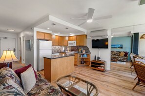 Una cocina moderna con armarios de madera, un microondas y un horno empotrado. Hay un comedor con una mesa de vidrio y sillas, y una sala de estar con un sofá y televisión.