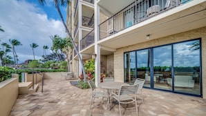 A multi-story building with a patio area featuring a stone floor, outdoor furniture, and a view of the surrounding landscape. - Kihei Beach #404- Serenity by the Shore (Kihei, Maui)