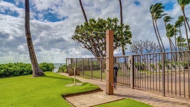 A gated entrance with a pathway leading to a seating area, palm trees, and a view of the ocean. - Kihei Beach #404- Serenity by the Shore (Kihei, Maui)