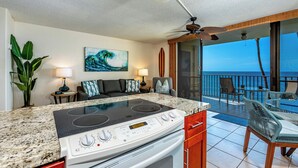 A kitchen with a stove, a dining area with chairs and a table, a ceiling fan, and a view of the ocean. - Kihei Beach #404- Serenity by the Shore (Kihei, Maui)