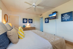 A bedroom with a bed, a ceiling fan, a wooden dresser, and wall art. - Kihei Beach #404- Serenity by the Shore (Kihei, Maui)