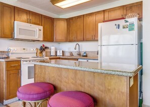 A kitchen with wooden cabinets, a white refrigerator, a microwave, a stove, and a sink. There are two red stools at the island.