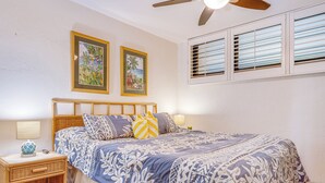 Une chambre à coucher comprenant un lit, deux tables de chevet, un ventilateur de plafond et deux fenêtres équipées de stores.