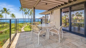 A covered outdoor patio with a table and chairs, overlooking a scenic view of the ocean and palm trees. - Oceanfront Gem with Mesmerizing Views and Lush Surroundings – Wailea Elua #1404 (Wailea, Maui)