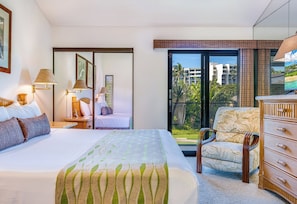 Habitación de hotel con una cama grande, una silla, un tocador y un balcón con vistas a edificios y palmeras.