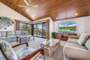A spacious living room with a ceiling fan, a large window with a view of palm trees, and a flat-screen TV.