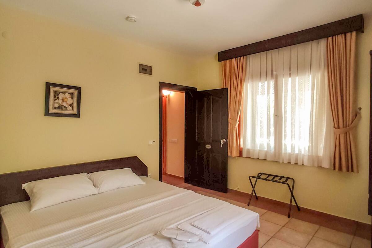 Une chambre d’hôtel avec un grand lit, une table de chevet, une petite chaise, une fenêtre avec des rideaux et une porte.