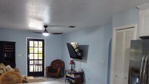A living room with a dining area, a TV mounted on the wall, a ceiling fan, and a refrigerator. - Great location.  Ten minutes from  Beach.  Peaceful retreat! (Merritt Island)