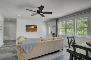 Un salón moderno con un sofá beige, un ventilador de techo, un cuadro de una puesta de sol y grandes ventanales con vistas a zonas verdes.