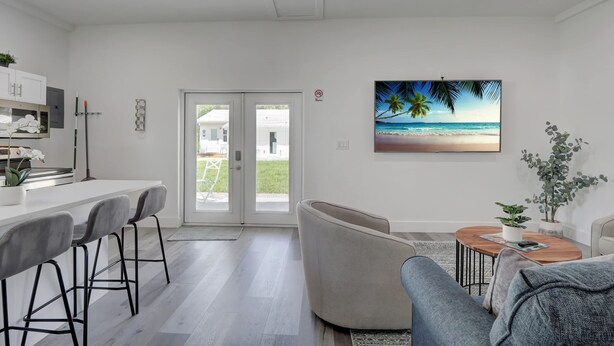 A modern living room with a large window, a dining area with bar stools, and a painting of a beach scene.