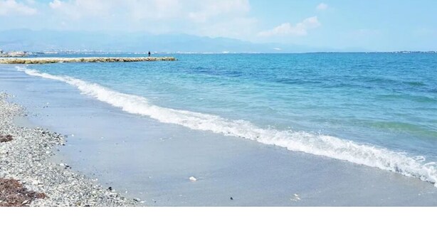 A beach with clear blue water, pebbles, and a distant rocky outcrop. - Gated Beachfront House - 10 Minutes from Kingston (Portmore)