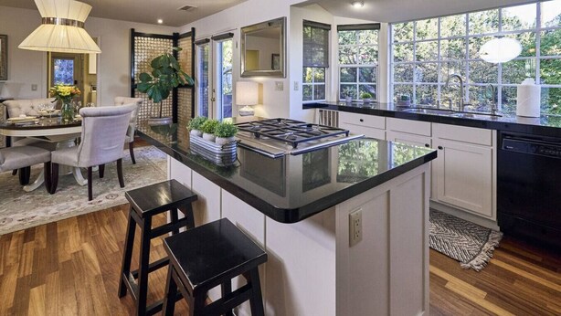 Una cocina moderna con una isla central, taburetes de bar, un comedor con candelabro y ventanas amplias que ofrecen vistas a zonas verdes.