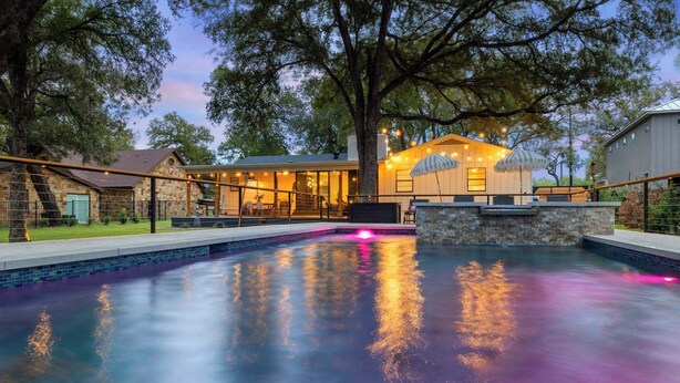 A swimming pool with a stone border, a covered patio area, and a large tree in the background. - The Creek Villa | Concierge Services (Horseshoe Bay)