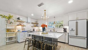 A modern kitchen with white cabinetry, a central island with bar stools, a built-in oven, and a refrigerator. - The Creek Villa | Concierge Services (Horseshoe Bay)