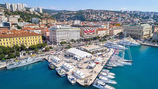Un port de plaisance où sont amarrés de nombreux yachts, un grand bâtiment avec une enseigne rouge, et une vue sur la ville en arrière-plan.