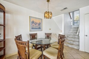 Un comedor con una mesa de vidrio rodeada de sillas de madera con asientos acolchados. Un candelabro cuelga del techo y un cuadro enmarcado está en la pared.