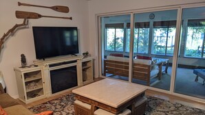 Ein Wohnzimmer mit Flachbildfernseher, Holzmöbeln, einem gemusterten Teppich und Blick durch große Glastüren.