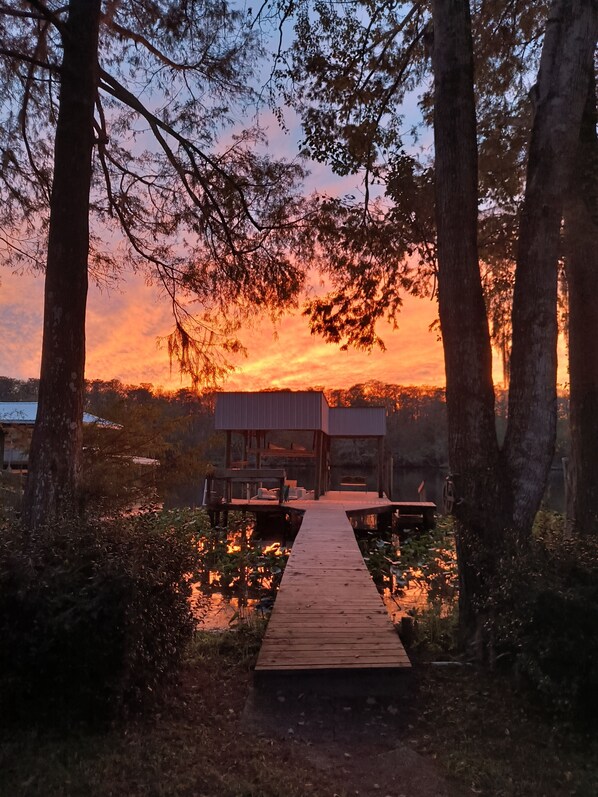 A wooden dock leading to a modern building with a flat roof, surrounded by trees and a sunset in the background.