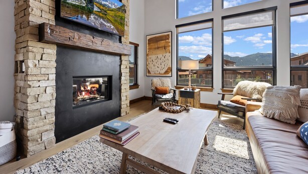 A cozy living room with a stone fireplace, large windows, and a comfortable seating area. - Estes Dream! End Unit with Patio, Indoor/Outdoor Fireplace and Jacuzzi (Estes Park)