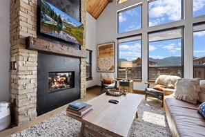 A cozy living room with a stone fireplace, large windows, and a comfortable seating area. - Estes Dream! End Unit with Patio, Indoor/Outdoor Fireplace and Jacuzzi (Estes Park)