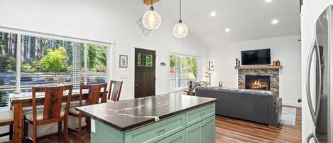 A spacious kitchen with a large island, wooden dining table, and chairs. There is a fireplace, a television, and a view of greenery outside.