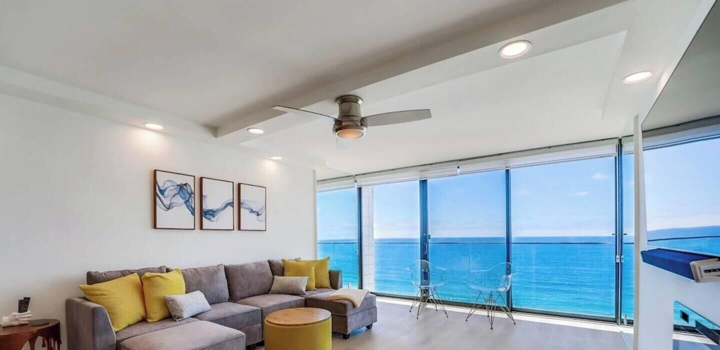 A modern living room with a large window overlooking the sea, a ceiling fan, and a sofa with cushions.
