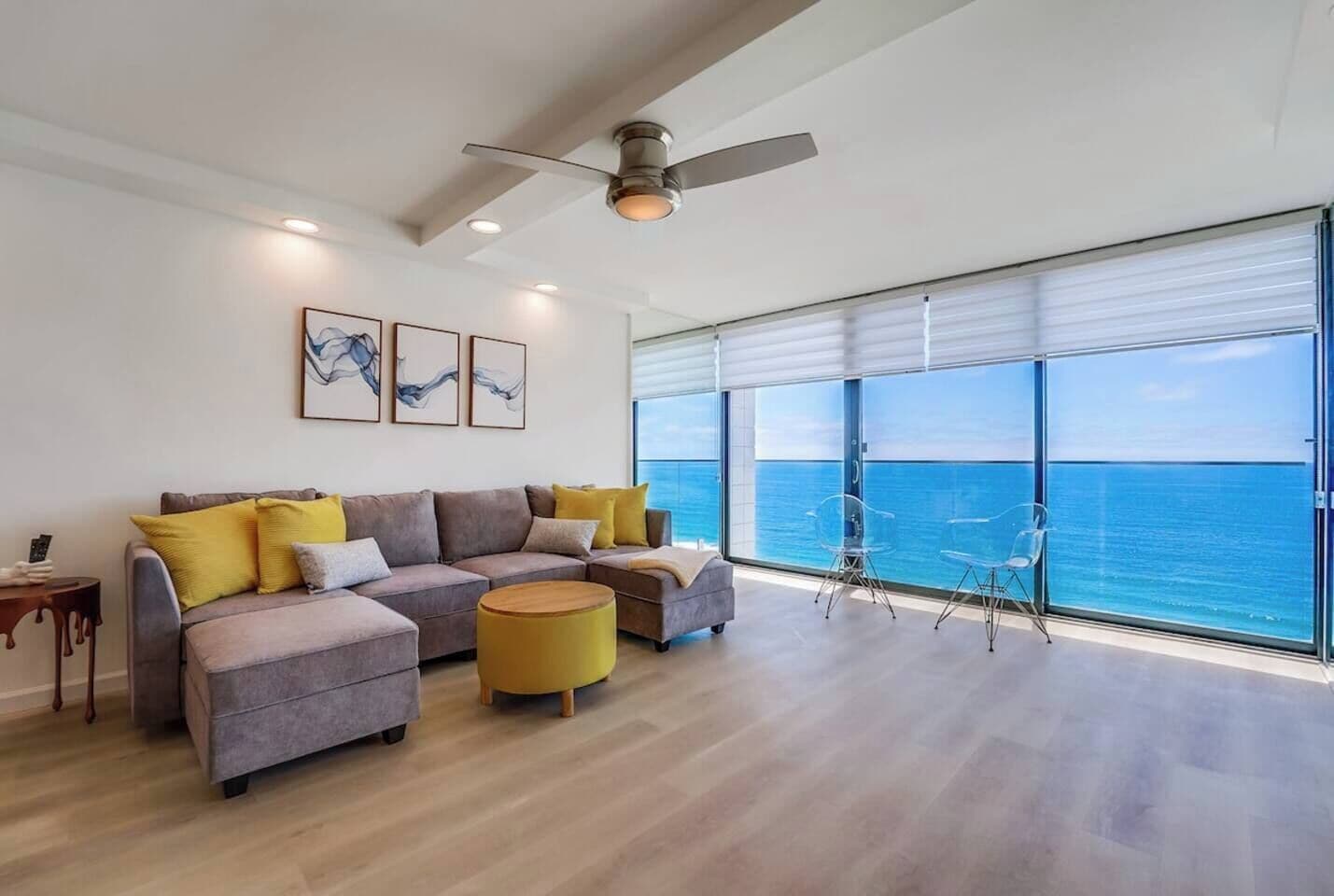 A modern living room with a sofa set, a round ottoman, and a view of the ocean through large windows.