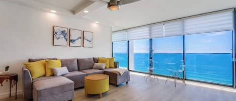 A modern living room with a sofa set, a round ottoman, and a view of the ocean through large windows.