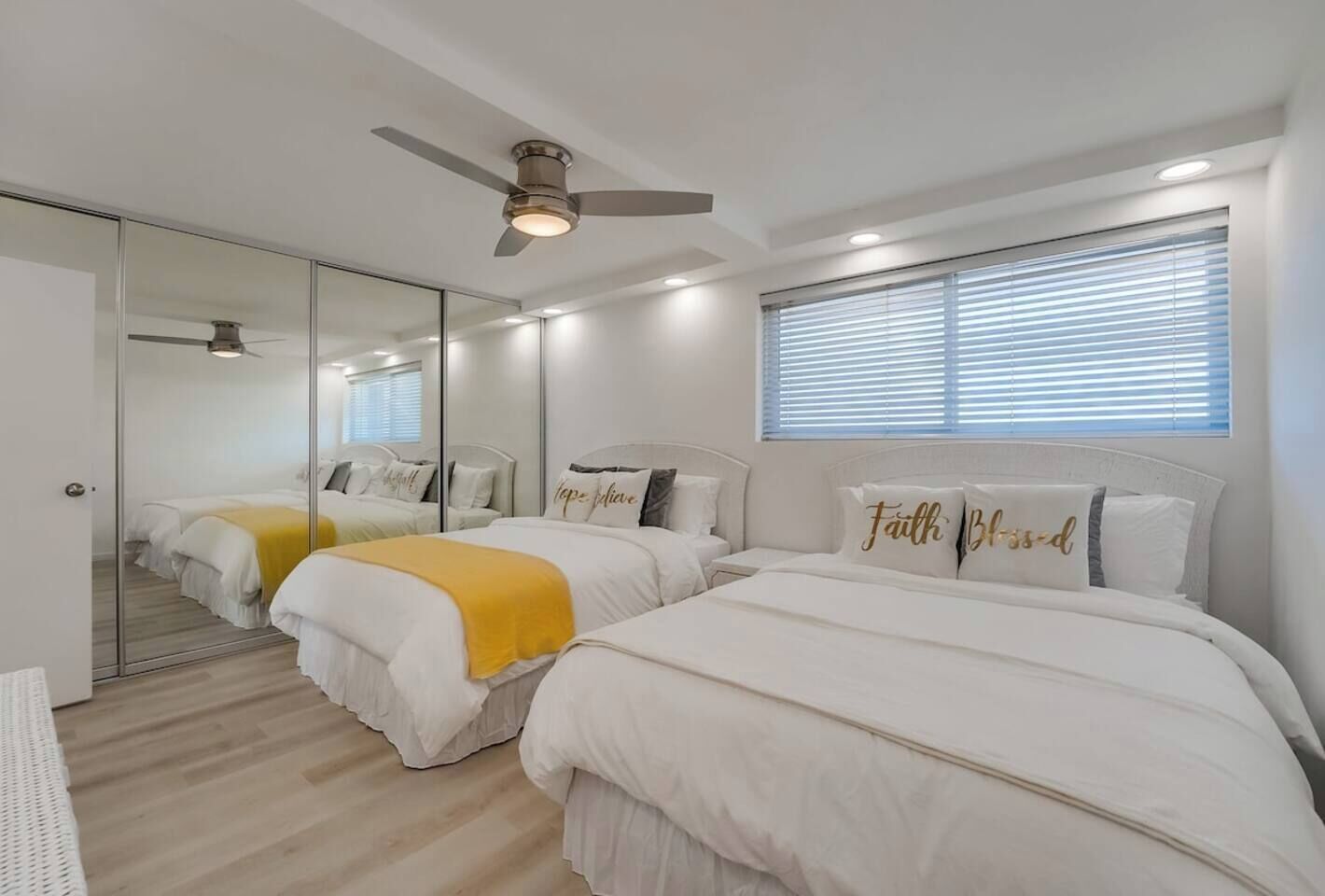 A hotel room with two beds, a ceiling fan, a large mirror, and a window with blinds.
