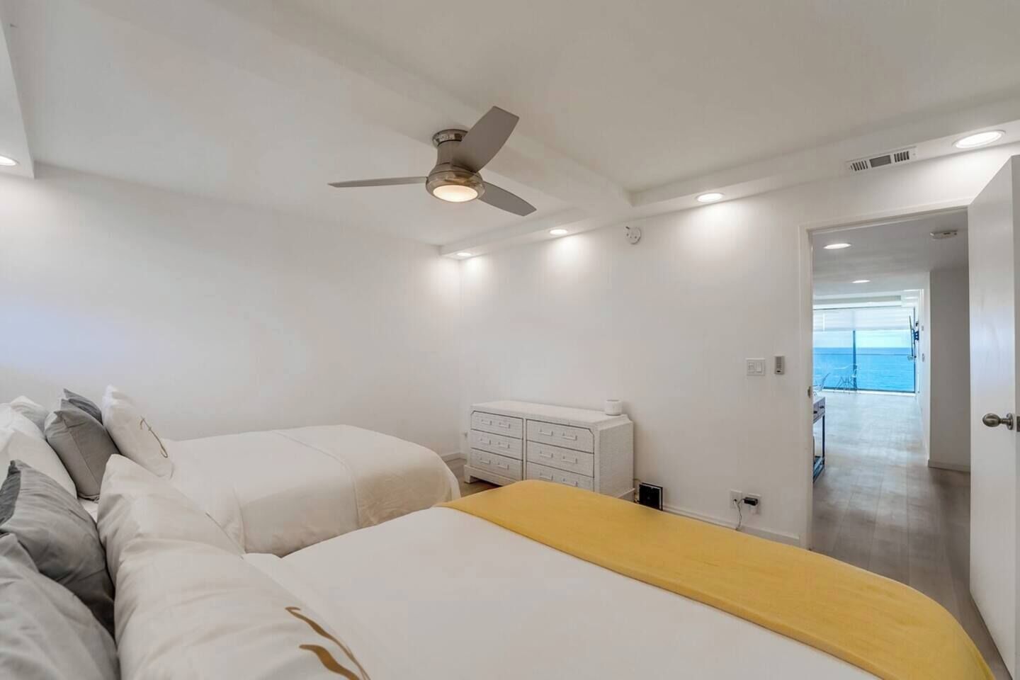 A bedroom with two beds, a ceiling fan, a dresser, and a view of the sea through an open door.