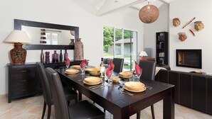 A dining area with a dark wooden table set for four, surrounded by black chairs. A large mirror hangs on the wall above the table, and there are decorative items like vases and a sun-shaped object on a dark wooden cabinet. - Les Villas de Fontenelles (Apremont)