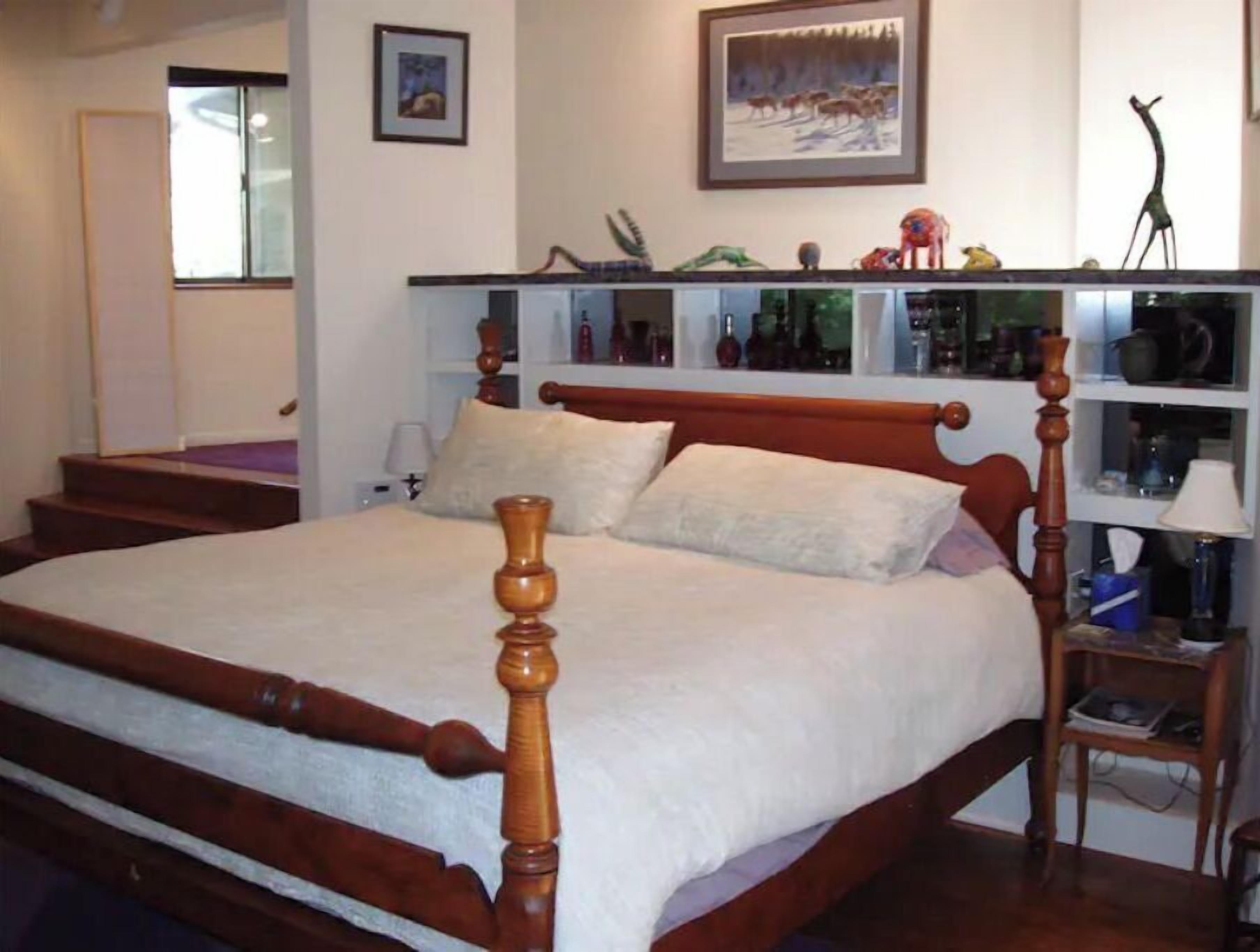 Une chambre à coucher avec un lit en bois, une table de chevet, une étagère contenant divers objets et une fenêtre donnant sur des arbres.