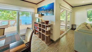 A room with a wooden dining table, chairs, a bookshelf, a painting, and a view of the ocean.