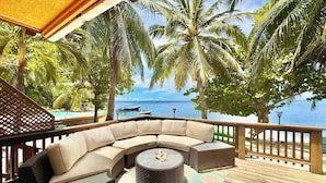 A wooden deck with a curved sofa, a round table, and a view of the ocean with palm trees.