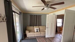 Quarto com sofá bege, ventilador de teto, porta e janela com cortinas.