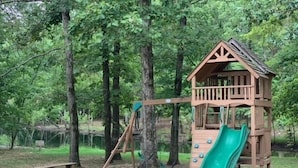 A wooden playground with a slide and climbing structure, surrounded by trees and a dirt ground.