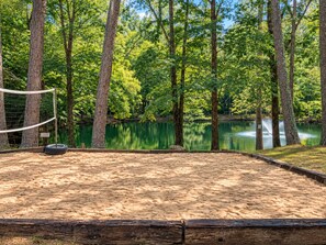 A sandy volleyball court with a net, surrounded by trees and a calm lake.