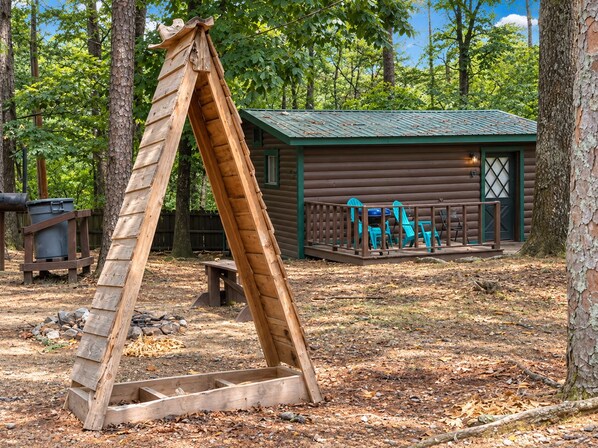 A wooden swing set in a wooded area with a cabin in the background.