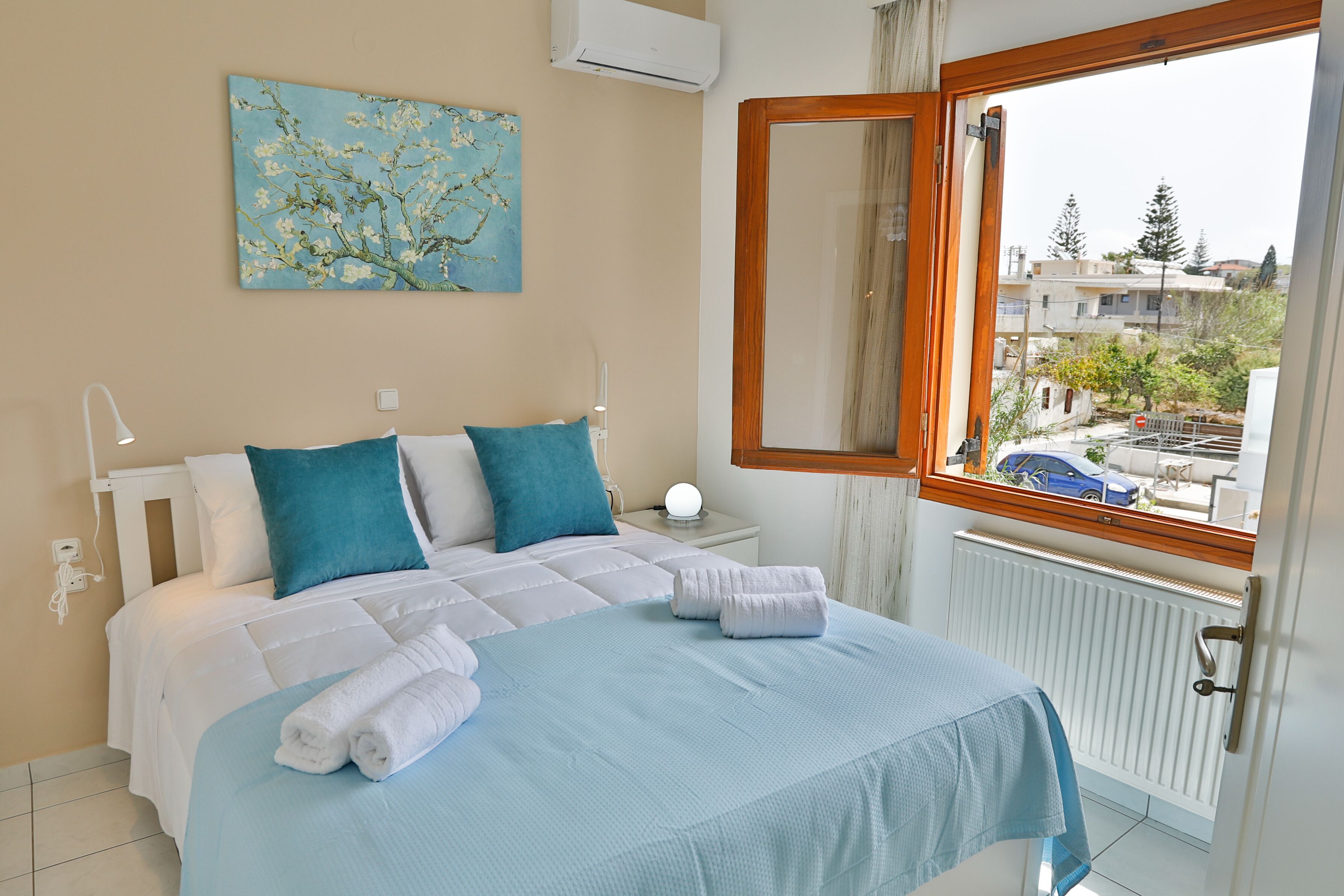Une chambre à coucher avec un lit, des oreillers, une table de chevet avec une lampe, un tableau au mur et une fenêtre donnant sur des bâtiments et des arbres.
