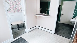 Tilava huone, jossa on vaalea puulattia, valkoinen ovi, pieni hylly koriste-esineitä varten ja peili seinällä.