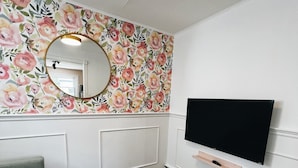 Huoneessa on kukallinen tapetti, pyöreä puinen pöytä, harmaa sohva ja seinälle kiinnitetty televisio.