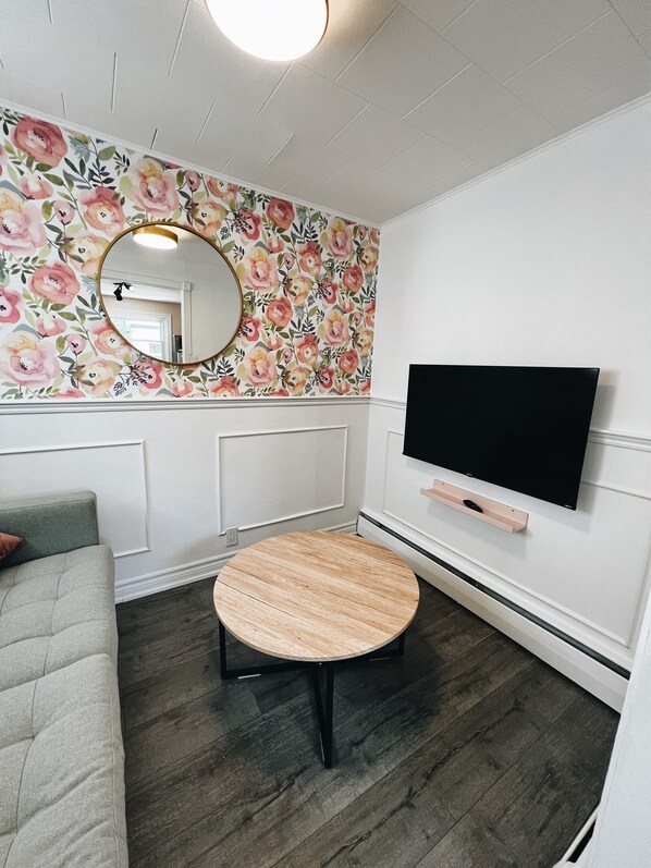 A room with a floral wallpaper, a round wooden table, a grey sofa, and a mounted TV.