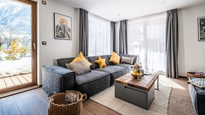 A modern living room with a grey sofa, a wooden coffee table, and a view of a snowy mountain landscape through large windows. - EYONG - Premium apartment with sauna and hot tub (Chamonix-Mont-Blanc)