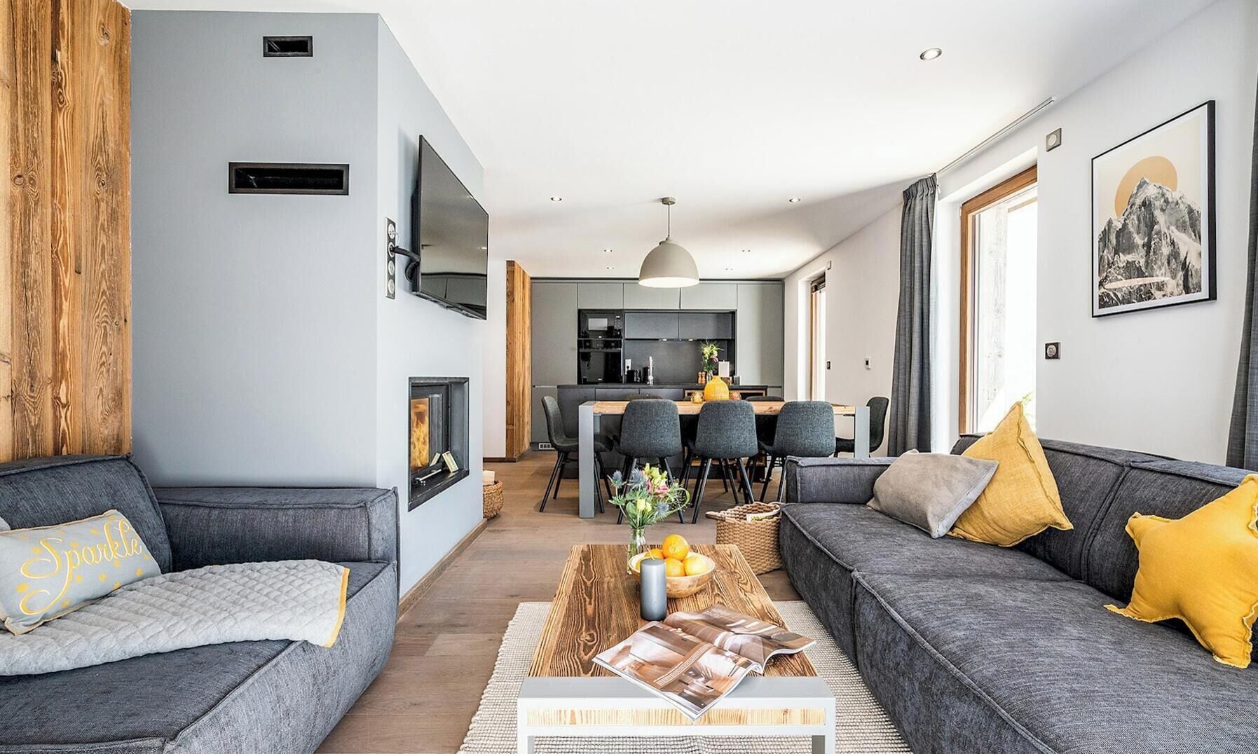 A modern living room with a fireplace, grey sofas, a dining area, and a kitchen in the background.