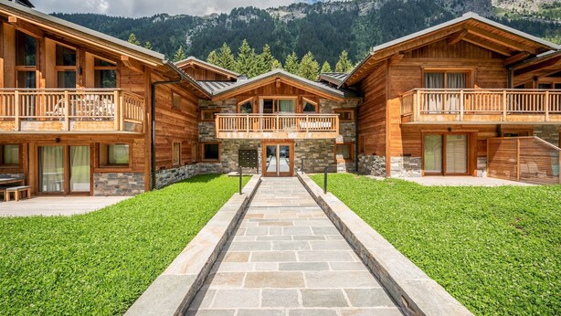 Una cabaña de madera con entrada central, sendero de piedra y balcones con vistas a un paisaje montañoso.